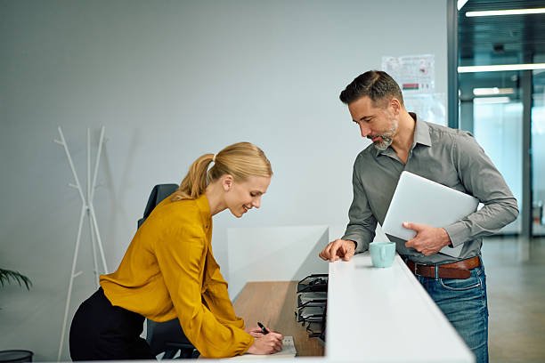 Businesswoman assisting a male client signing a registration form at a modern office reception area, showing professional interaction and welcoming atmosphere